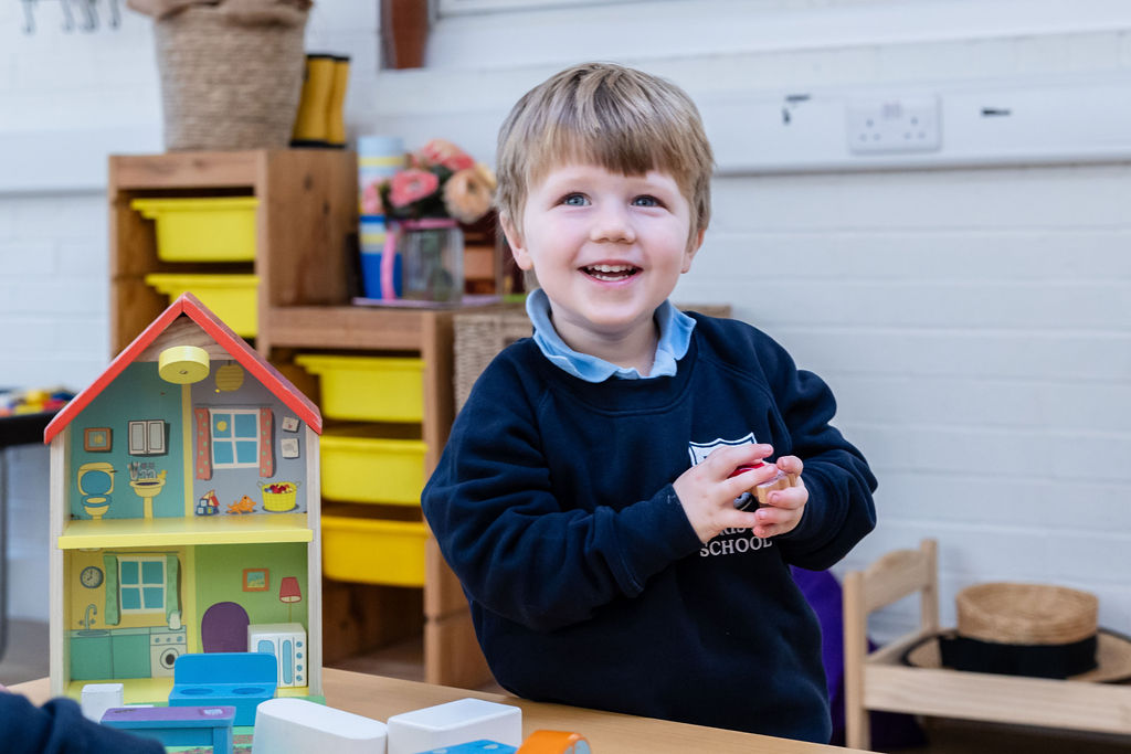 A boy in Horris Hill Nursery uniform stands by a table holding a toy and smiling happily
